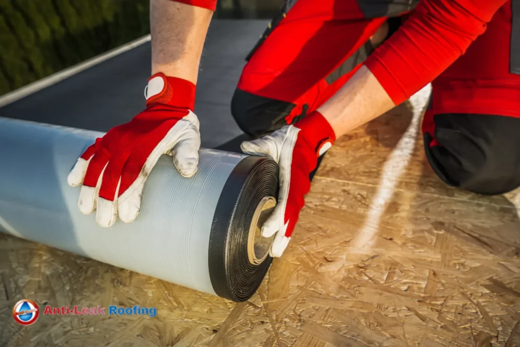 A worker in red overalls applies a waterproof sealing tape to a roof, wearing gloves and kneeling on the surface.