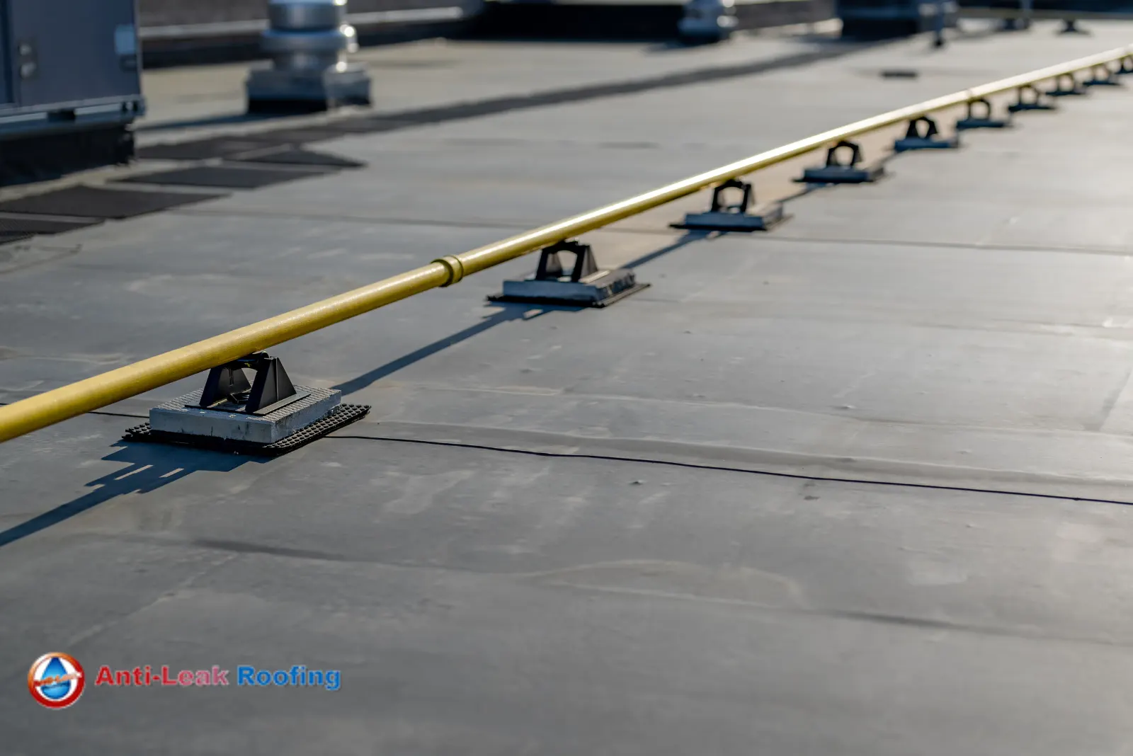 A flat rooftop with a yellow safety guardrail. The rooftop surface is composed of large panels. A logo for "Anti-Leak Roofing" is visible in the bottom left corner.