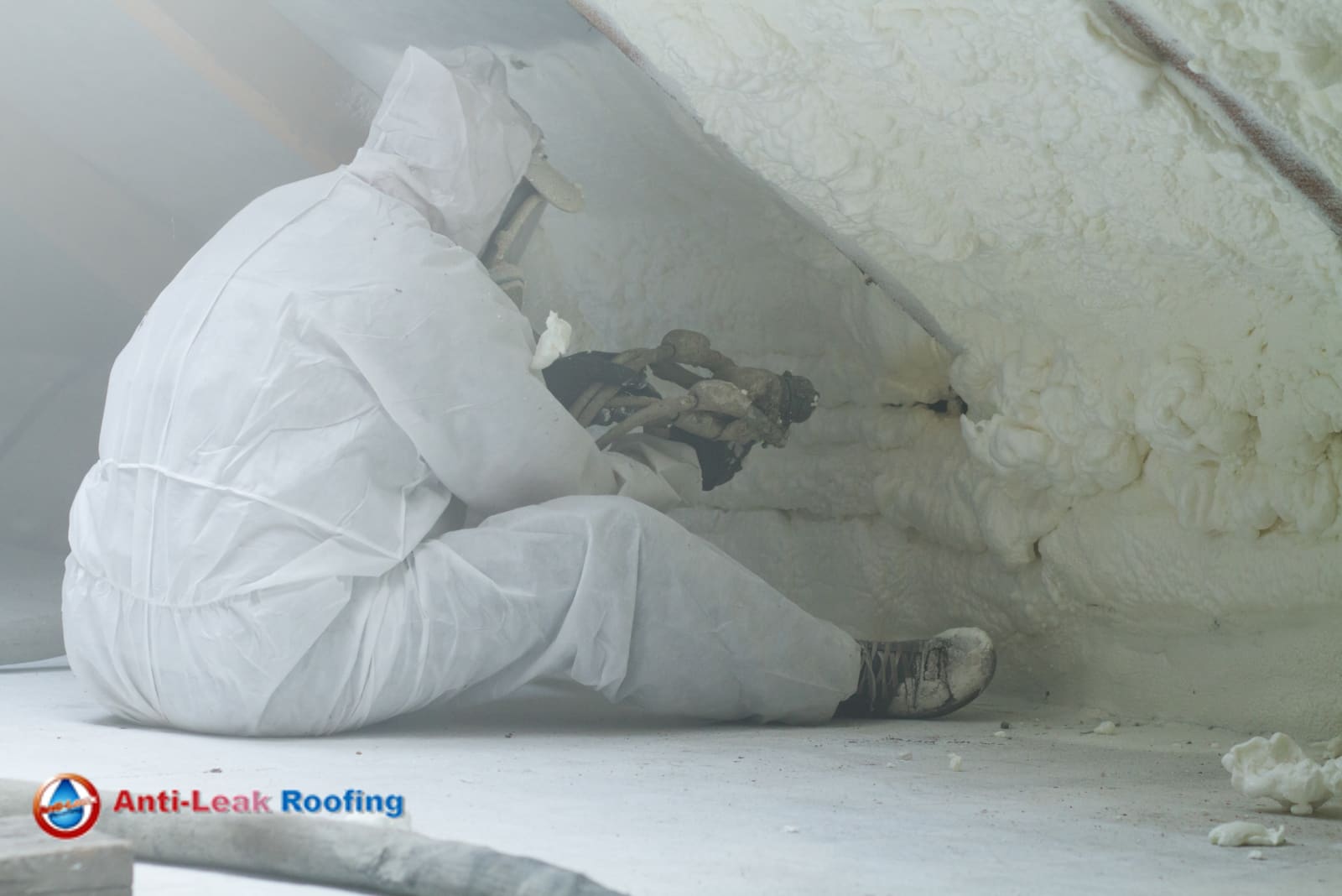 A worker in white protective gear applies foam insulation to an attic space. The "Anti-Leak Roofing" logo is visible in the lower-left corner.