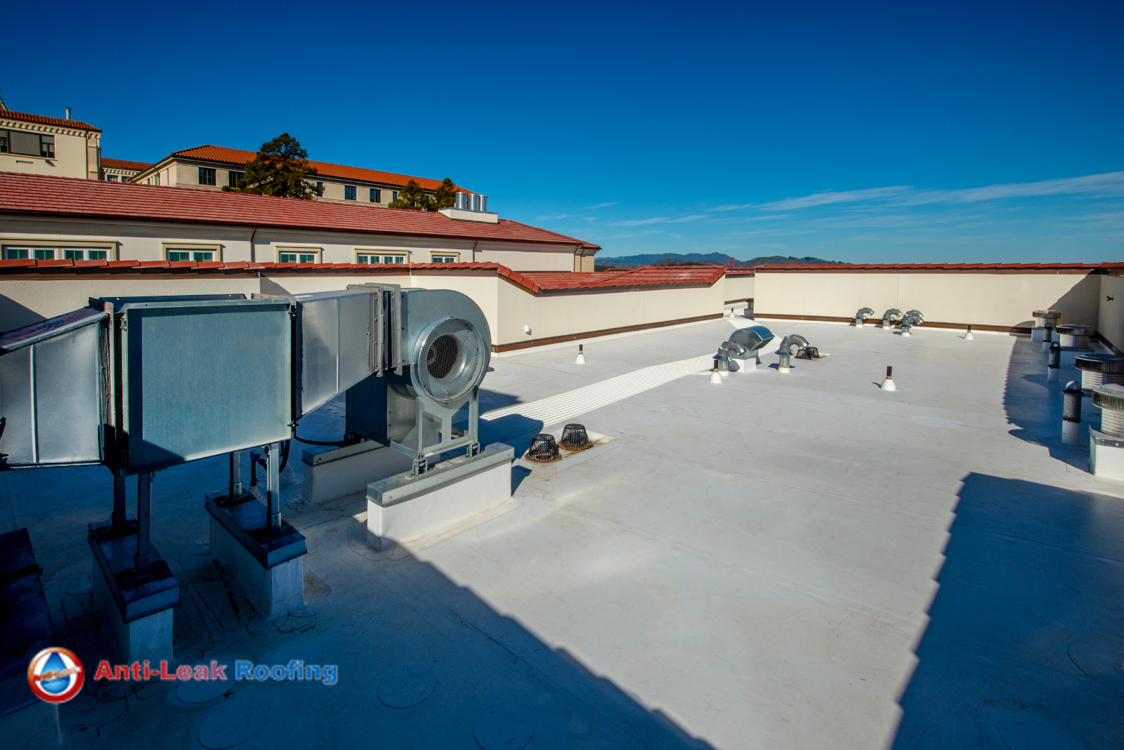 Rooftop view of a building with HVAC units and vents on a flat, white surface. Buildings with red-tiled roofs can be seen in the background. A logo for "Anti-Leak Roofing" is in the bottom left corner.